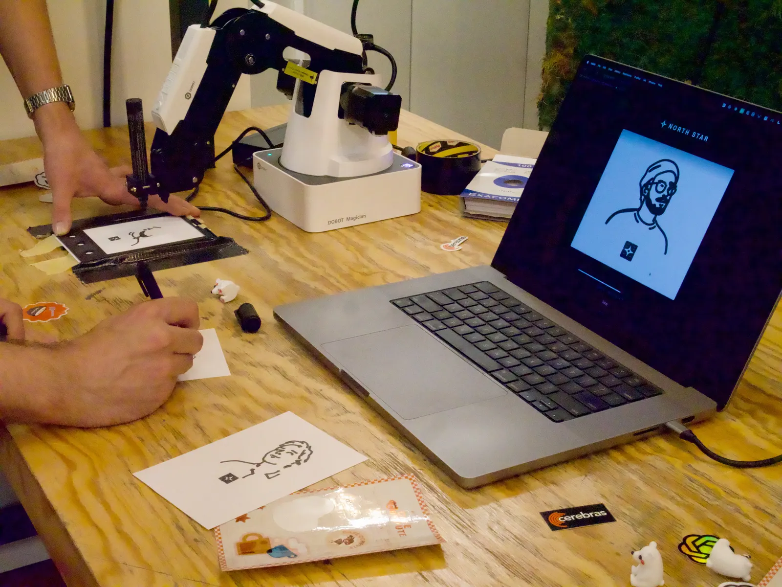 MARK-E tekent een portret op een kaartje; naast de robot staat een laptop met de gegenereerde lijntekening